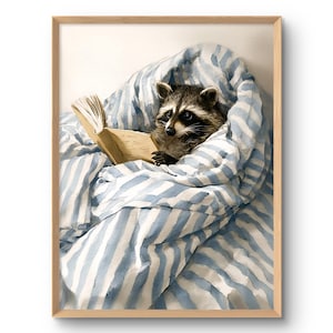 May include: A framed watercolor print depicts a raccoon reading a book while nestled in a blue and white striped blanket. The raccoon is brown and black with a white face, and the book is open with visible pages.
