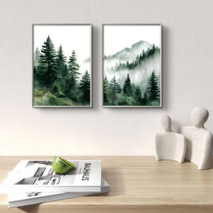 May include: Two framed watercolor paintings depict forest scenes. One shows tall evergreen trees, the other a mountain range with trees in the mist. The paintings have silver frames and are displayed on a white wall.