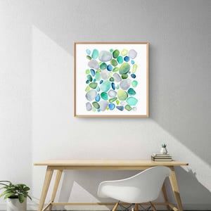 Puede incluir: Una impresi&oacute;n en acuarela enmarcada con un dise&ntilde;o abstracto de formas redondeadas en tonos de azul, verde y gris. La obra de arte se exhibe en una pared blanca sobre un escritorio de madera y una silla blanca.
