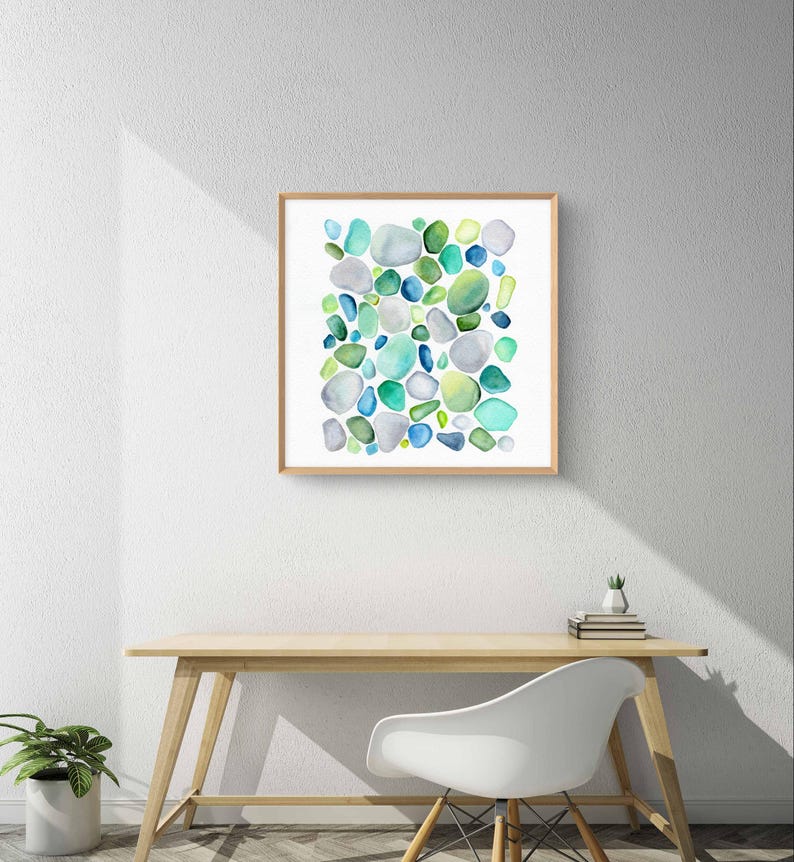 Puede incluir: Una impresi&oacute;n en acuarela enmarcada con un dise&ntilde;o abstracto de formas redondeadas en tonos de azul, verde y gris. La obra de arte se exhibe en una pared blanca sobre un escritorio de madera y una silla blanca.