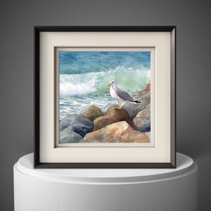 May include: Watercolor painting of a seagull perched on rocks near the ocean. The artwork features a white and gray seagull with a yellow beak and legs. The background shows a blue ocean with a breaking wave. The painting is framed in a black frame.