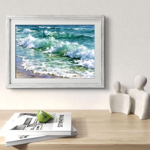 May include: A framed landscape painting of ocean waves crashing onto a sandy shore, featuring shades of deep blue, turquoise, and white sea foam. The artwork is displayed in a light grey frame, positioned above a stack of books and two white figures.