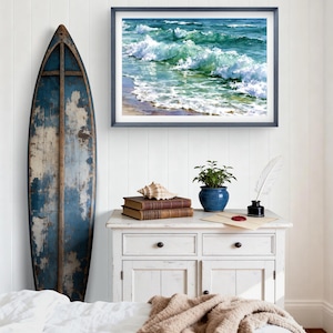 May include: A framed horizontal wall art print depicting ocean waves crashing on a sandy shore, displayed above a white wooden cabinet featuring two stacked books, a seashell, a potted green plant, and a white quill pen in an inkwell.
