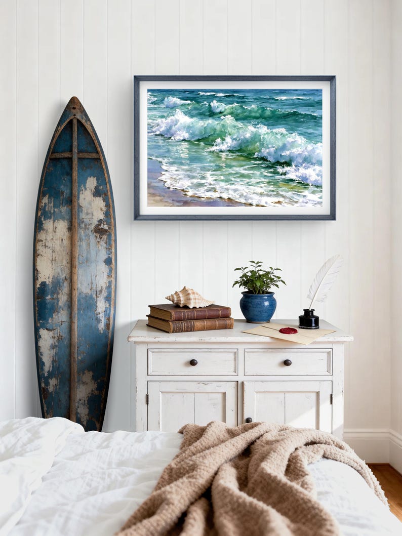 May include: A framed horizontal wall art print depicting ocean waves crashing on a sandy shore, displayed above a white wooden cabinet featuring two stacked books, a seashell, a potted green plant, and a white quill pen in an inkwell.