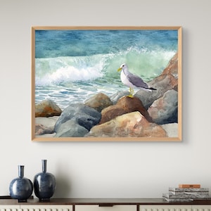 May include: Watercolor painting of a seagull perched on rocks at the edge of the ocean. The artwork features a light brown wooden frame, with the bird in the center, and a wave crashing in the background. The color palette includes shades of blue, gray, and brown.