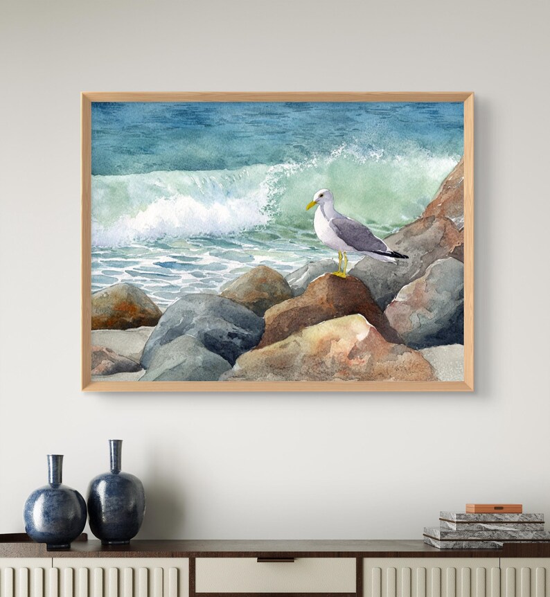 May include: Watercolor painting of a seagull perched on rocks at the edge of the ocean. The artwork features a light brown wooden frame, with the bird in the center, and a wave crashing in the background. The color palette includes shades of blue, gray, and brown.