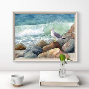 May include: Watercolor painting of a seagull perched on rocks near the ocean. The artwork features a white-framed print with a blue and green ocean wave in the background. The seagull has gray wings and a yellow beak.