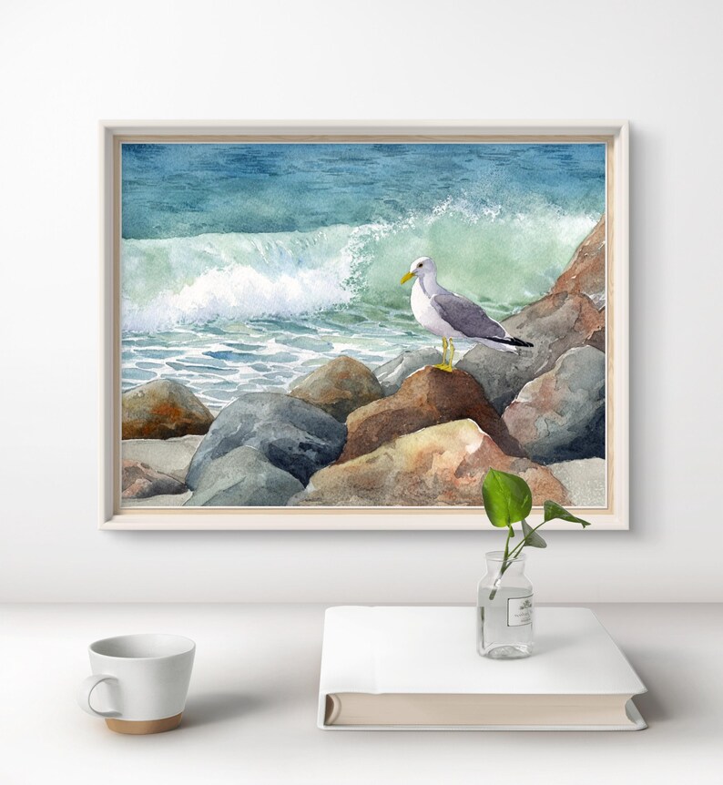 May include: Watercolor painting of a seagull perched on rocks near the ocean. The artwork features a white-framed print with a blue and green ocean wave in the background. The seagull has gray wings and a yellow beak.