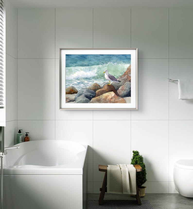 May include: A framed watercolor painting of a seagull perched on rocks near the ocean. The artwork features a crashing wave with shades of blue and green. The rocks are brown and gray, and the seagull has gray and white feathers.