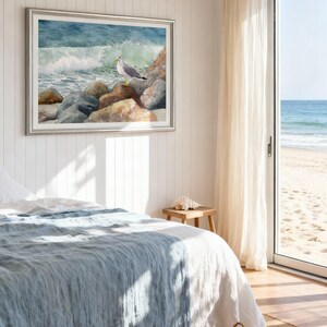May include: A framed watercolor painting of a seagull perched on rocks near the ocean. The artwork is in a silver frame and hangs on a white paneled wall. A bed with a blue comforter and a woven basket are in the foreground.