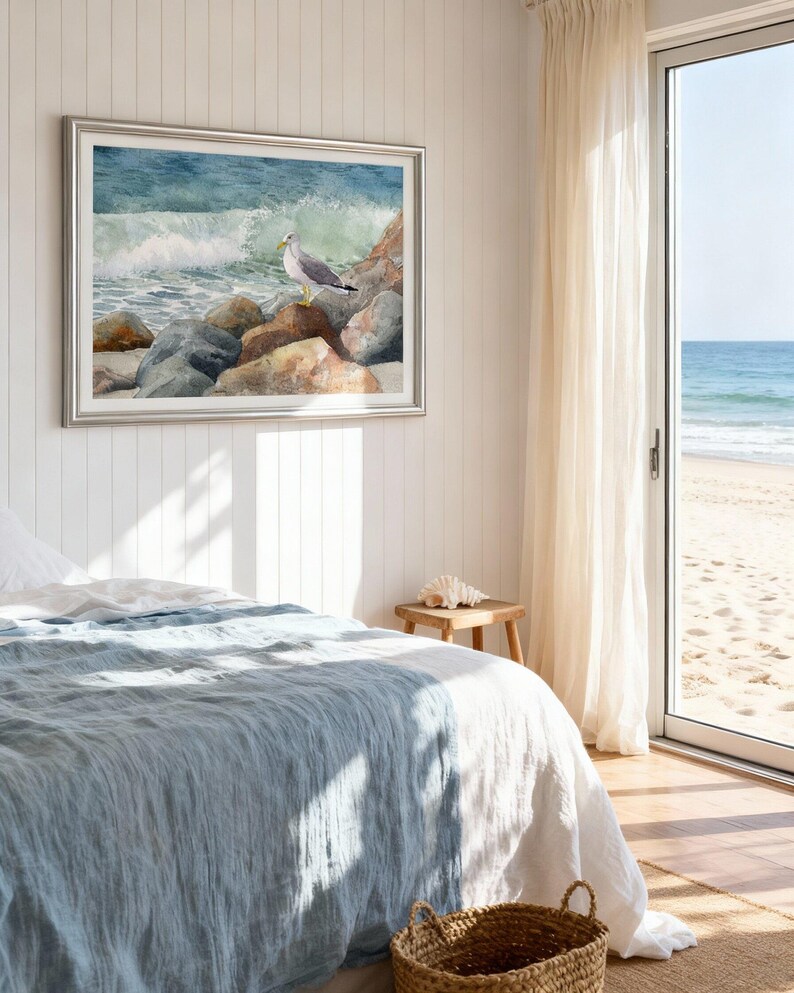 May include: A framed watercolor painting of a seagull perched on rocks near the ocean. The artwork is in a silver frame and hangs on a white paneled wall. A bed with a blue comforter and a woven basket are in the foreground.