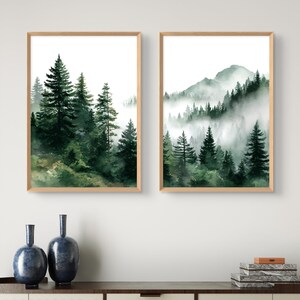 May include: Two framed watercolor prints depict a misty forest scene with tall evergreen trees. The artwork features shades of green and brown, with a light wooden frame. The prints are displayed above a dark wooden surface.