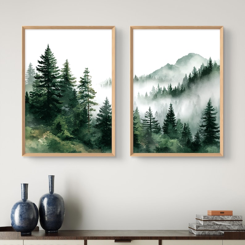 May include: Two framed watercolor prints depict a misty forest scene with tall evergreen trees. The artwork features shades of green and brown, with a light wooden frame. The prints are displayed above a dark wooden surface.