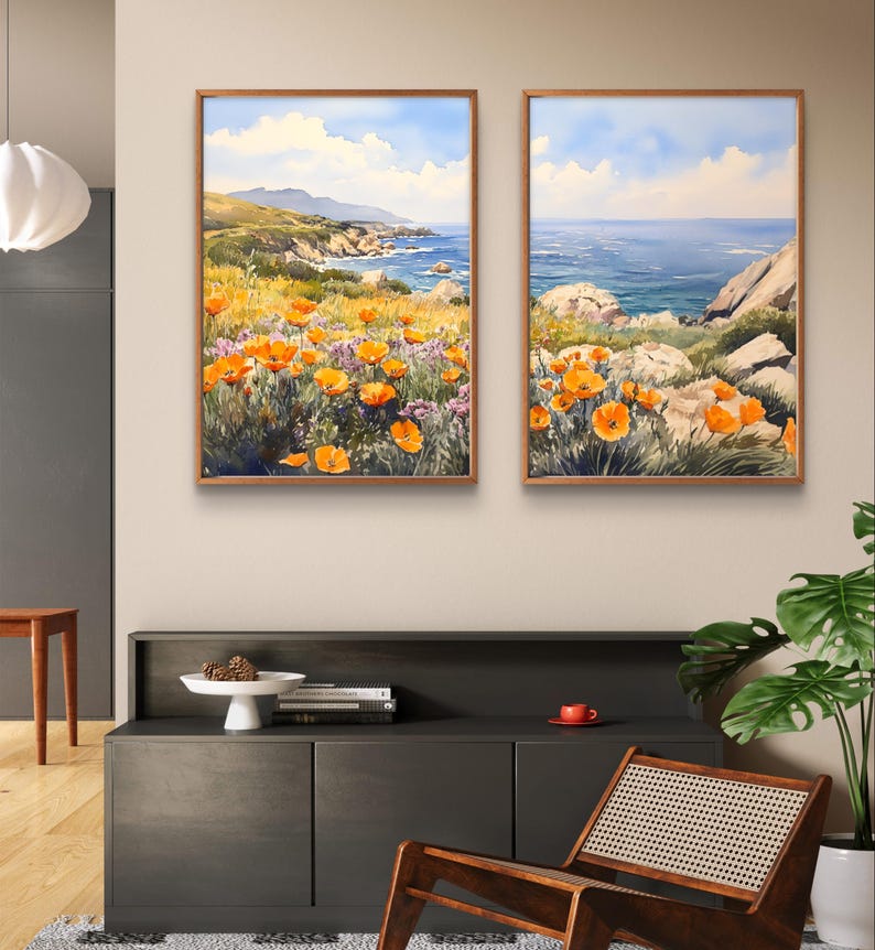 May include: Two framed watercolor paintings depicting a coastal landscape with orange wildflowers in the foreground and a blue ocean in the background. The paintings are hung on a wall above a dark grey cabinet.