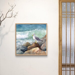 May include: Watercolor painting of a seagull perched on rocks, with a crashing wave in the background. The artwork is framed in a light brown wooden frame. The color palette includes shades of blue, gray, brown, and white.