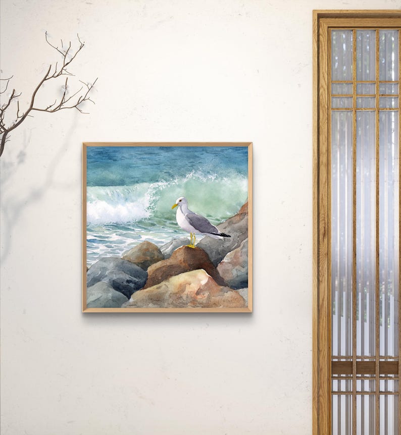 May include: Watercolor painting of a seagull perched on rocks, with a crashing wave in the background. The artwork is framed in a light brown wooden frame. The color palette includes shades of blue, gray, brown, and white.