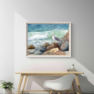 May include: Watercolor painting of a seagull perched on rocks near the ocean. The artwork features a white-framed print with a light blue and green wave crashing in the background. The seagull has gray and white feathers with a yellow beak.