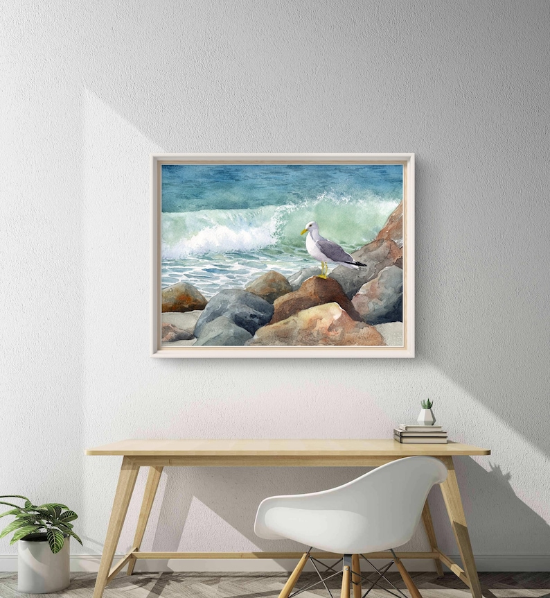 May include: Watercolor painting of a seagull perched on rocks near the ocean. The artwork features a white-framed print with a light blue and green wave crashing in the background. The seagull has gray and white feathers with a yellow beak.