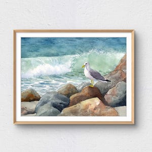 May include: Watercolor painting of a seagull perched on rocks at the edge of the ocean. The artwork features a light brown wooden frame, a blue-green ocean with a breaking wave, and a gray and white seagull with yellow legs and beak.