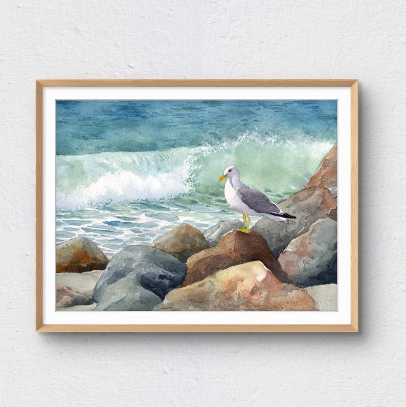 May include: Watercolor painting of a seagull perched on rocks at the edge of the ocean. The artwork features a light brown wooden frame, a blue-green ocean with a breaking wave, and a gray and white seagull with yellow legs and beak.