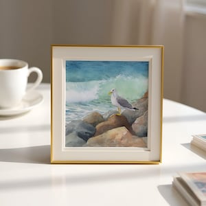 May include: A framed watercolor painting of a seagull perched on rocks, with a crashing wave in the background. The artwork is set in a gold frame and rests on a white surface. A cup of coffee is visible in the background.