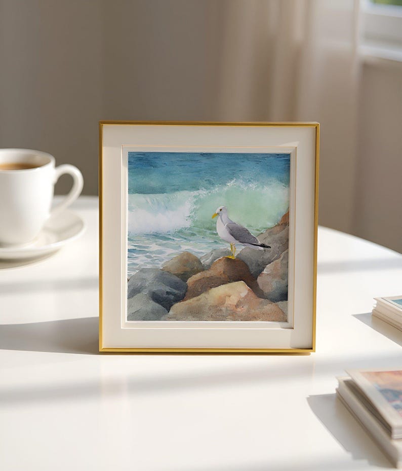 May include: A framed watercolor painting of a seagull perched on rocks, with a crashing wave in the background. The artwork is set in a gold frame and rests on a white surface. A cup of coffee is visible in the background.