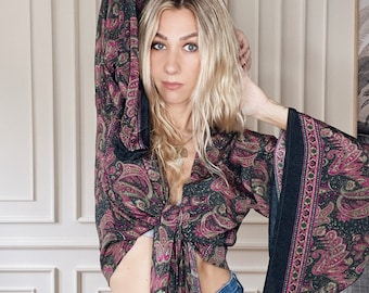 Puede incluir: Una mujer lleva una blusa corta de color negro y rosa con estampado de paisley, mangas largas y un nudo en la parte delantera. La blusa tiene un escote en V profundo y está anudada a la cintura. La mujer lleva pantalones vaqueros azules.