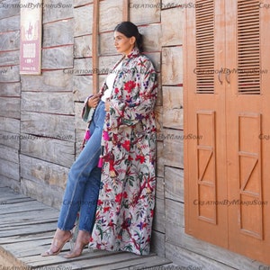 May include: A long, white floral print robe with pink, red, and blue accents. The robe has a tassel detail on the sleeves and a long, flowing silhouette. The model is wearing the robe with blue jeans and heels.