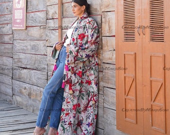 May include: A long, white floral print robe with pink, red, and blue accents. The robe has a tassel detail on the sleeves and a long, flowing silhouette. The model is wearing the robe with blue jeans and heels.