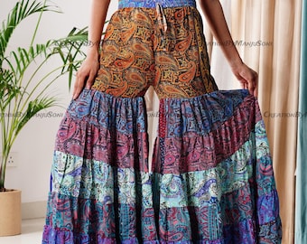Puede incluir: Pantalones palazzo con estampado de cachemira en tonos naranja, azul, morado y rosa. Los pantalones tienen cintura con cordón y perneras con volantes escalonados. La tela parece ligera y fluida, adecuada para climas cálidos.
