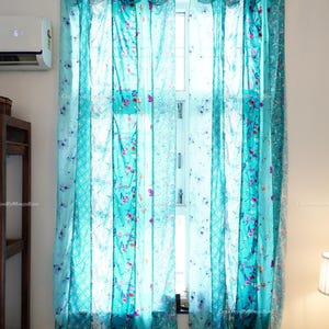 May include: A pair of sheer window curtains featuring a patchwork design of turquoise and teal fabric panels. The curtains are adorned with various floral and geometric patterns in shades of pink, white, and blue, and are hung from a rod with loops.
