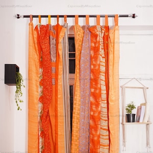 May include: Orange and peach colored fabric curtains with a window behind them. The curtains are made of multiple pieces of fabric sewn together. The curtains are hung on a black metal rod.