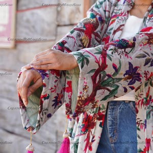 May include: A white kimono with a floral print featuring birds and flowers. The kimono has long sleeves with tassels at the ends. The kimono is worn over a white shirt and blue jeans.
