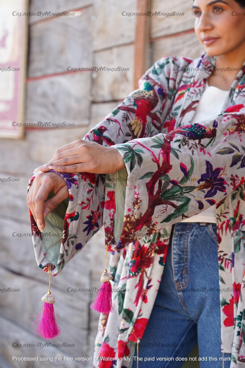 May include: A white kimono with a floral print featuring birds and flowers. The kimono has long sleeves with tassels at the ends. The kimono is worn over a white shirt and blue jeans.