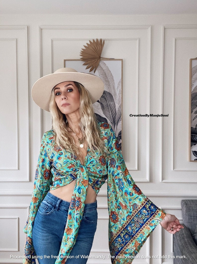 May include: A woman wearing a blue and green floral print blouse with a tie front and bell sleeves. She is also wearing a wide-brimmed hat and blue jeans.