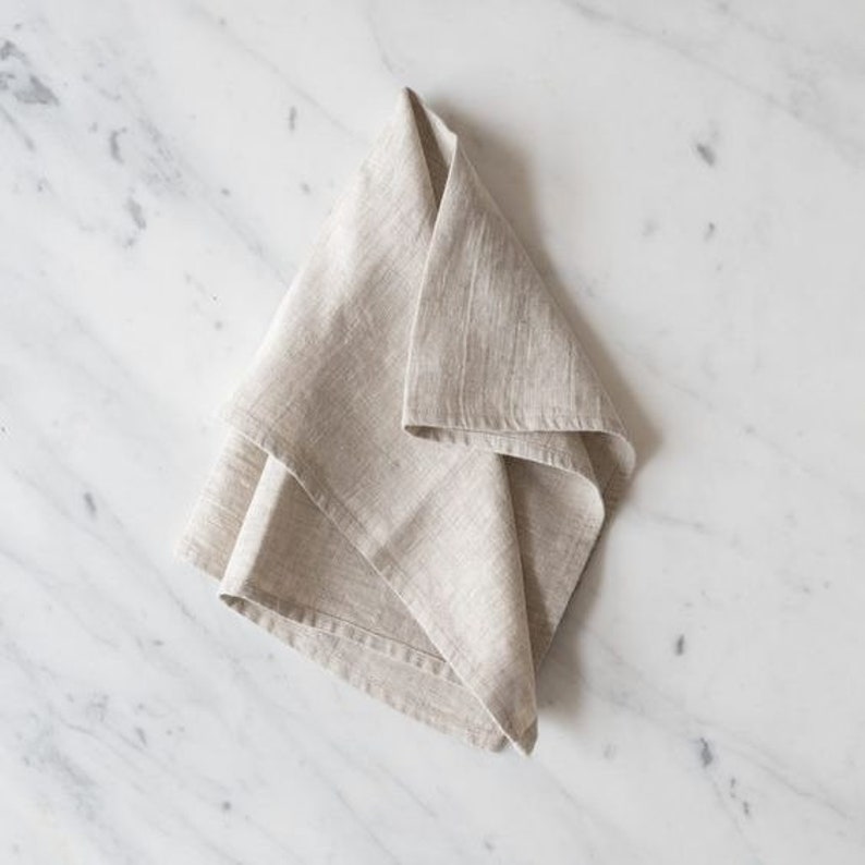 Pu&ograve; includere: Un asciugamano da cucina in lino piegato di colore beige chiaro. L'asciugamano &egrave; realizzato in tessuto naturale e ha una superficie leggermente strutturata.