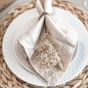 Pu&ograve; includere: Un tovagliolo di lino bianco con un sottopiatto in fibra naturale e un mazzo di fiori secchi. Il tovagliolo &egrave; piegato e legato con uno spago.