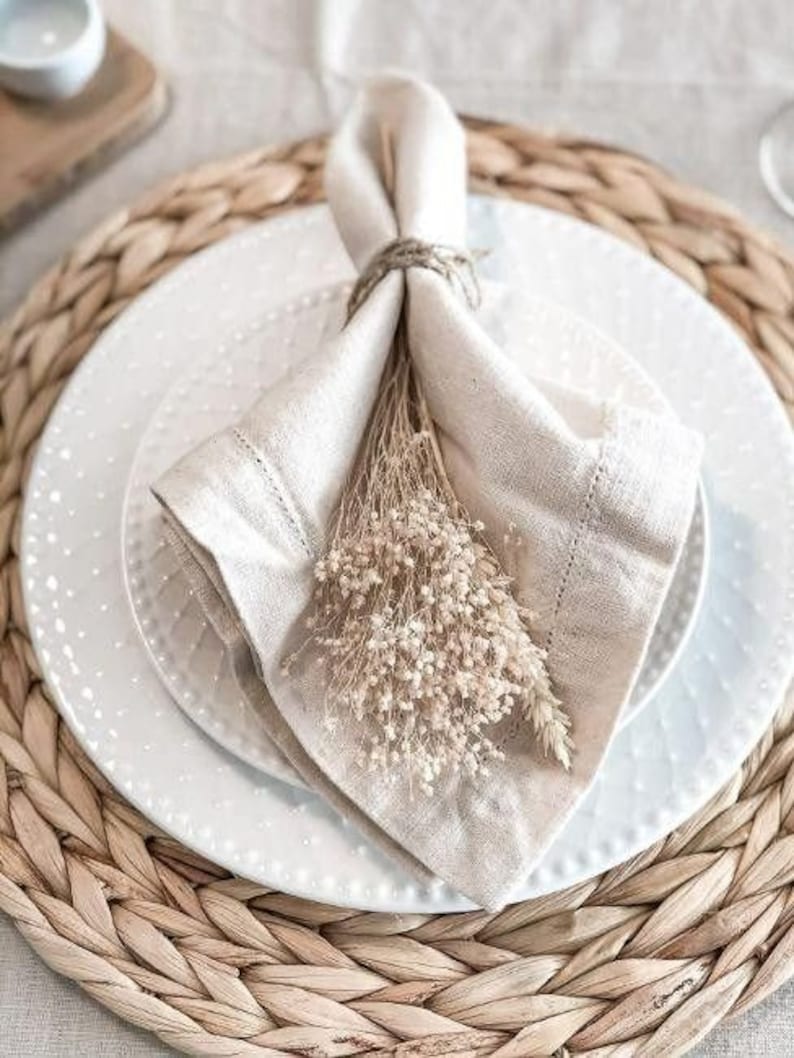 Pu&ograve; includere: Un tovagliolo di lino bianco con un sottopiatto in fibra naturale e un mazzo di fiori secchi. Il tovagliolo &egrave; piegato e legato con uno spago.