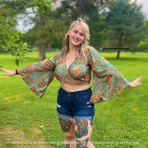 May include: A woman wearing a teal and red paisley print wrap top with bell sleeves. The top is cropped and shows a bit of her midriff. She is also wearing dark blue denim shorts with distressed details. She is standing in a grassy field with trees in the background.
