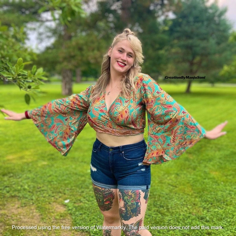 May include: A woman wearing a teal and red paisley print wrap top with bell sleeves. The top is cropped and shows a bit of her midriff. She is also wearing dark blue denim shorts with distressed details. She is standing in a grassy field with trees in the background.
