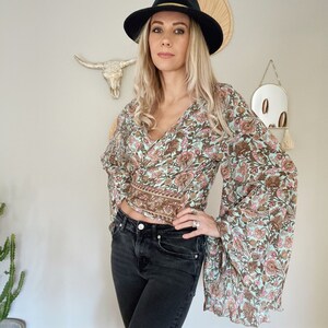 May include: A woman wearing a light blue floral print blouse with long bell sleeves and a black fedora hat. She is also wearing black jeans.
