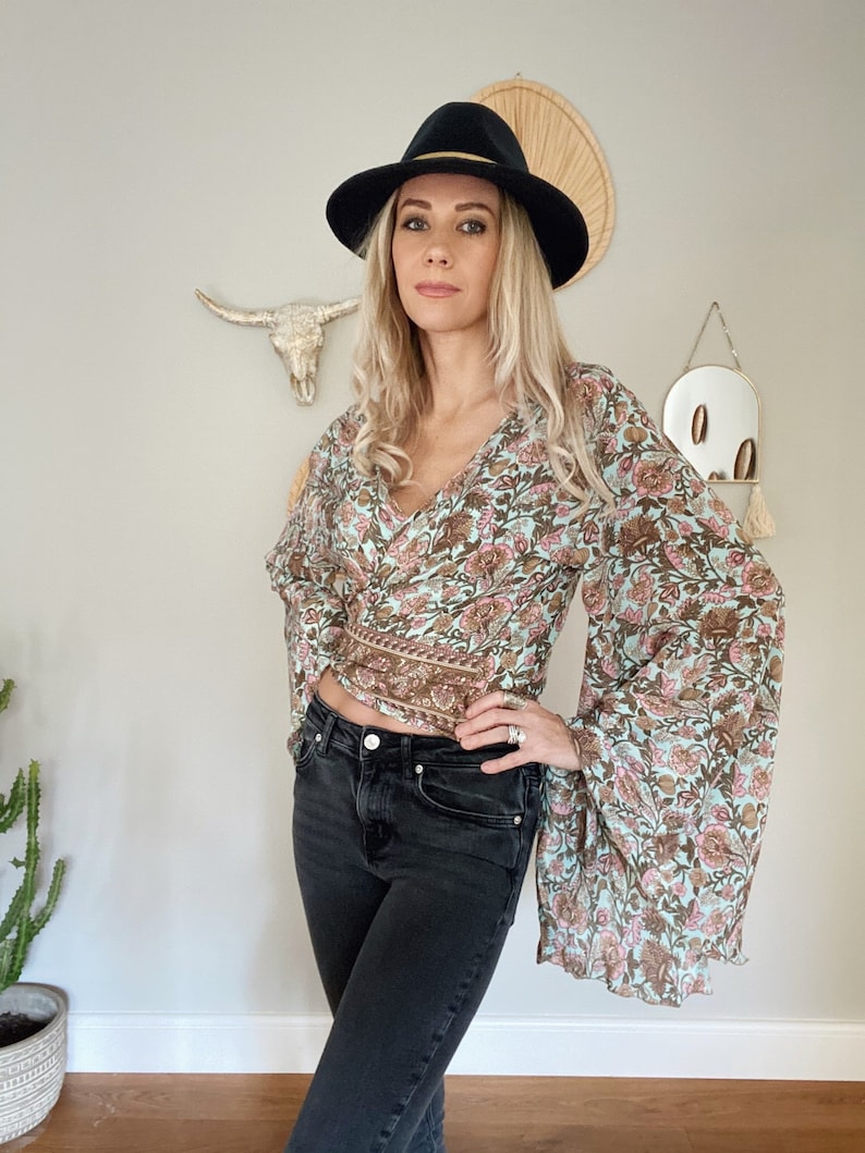 May include: A woman wearing a light blue floral print blouse with long bell sleeves and a black fedora hat. She is also wearing black jeans.