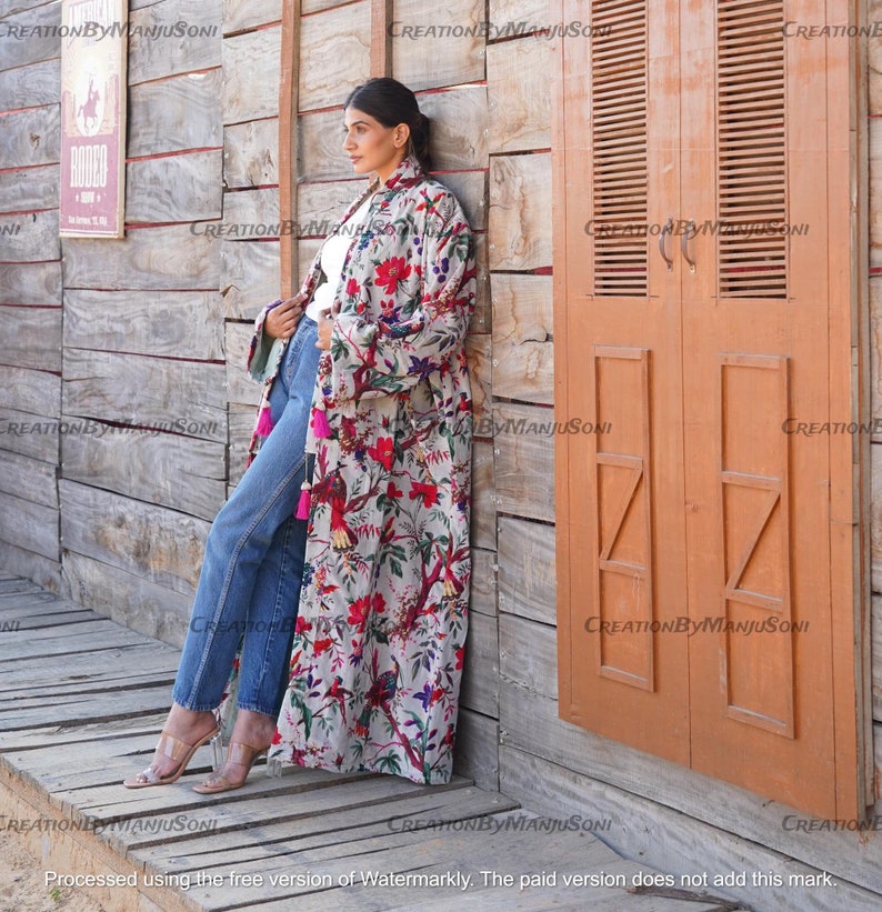 May include: A long, white floral print robe with pink, red, and blue accents. The robe has a tassel detail on the sleeves and a long, flowing silhouette. The model is wearing the robe with blue jeans and heels.