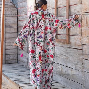 May include: A white long-sleeved robe with a floral print featuring colorful birds and flowers. The robe has a pink tassel on the sleeve.