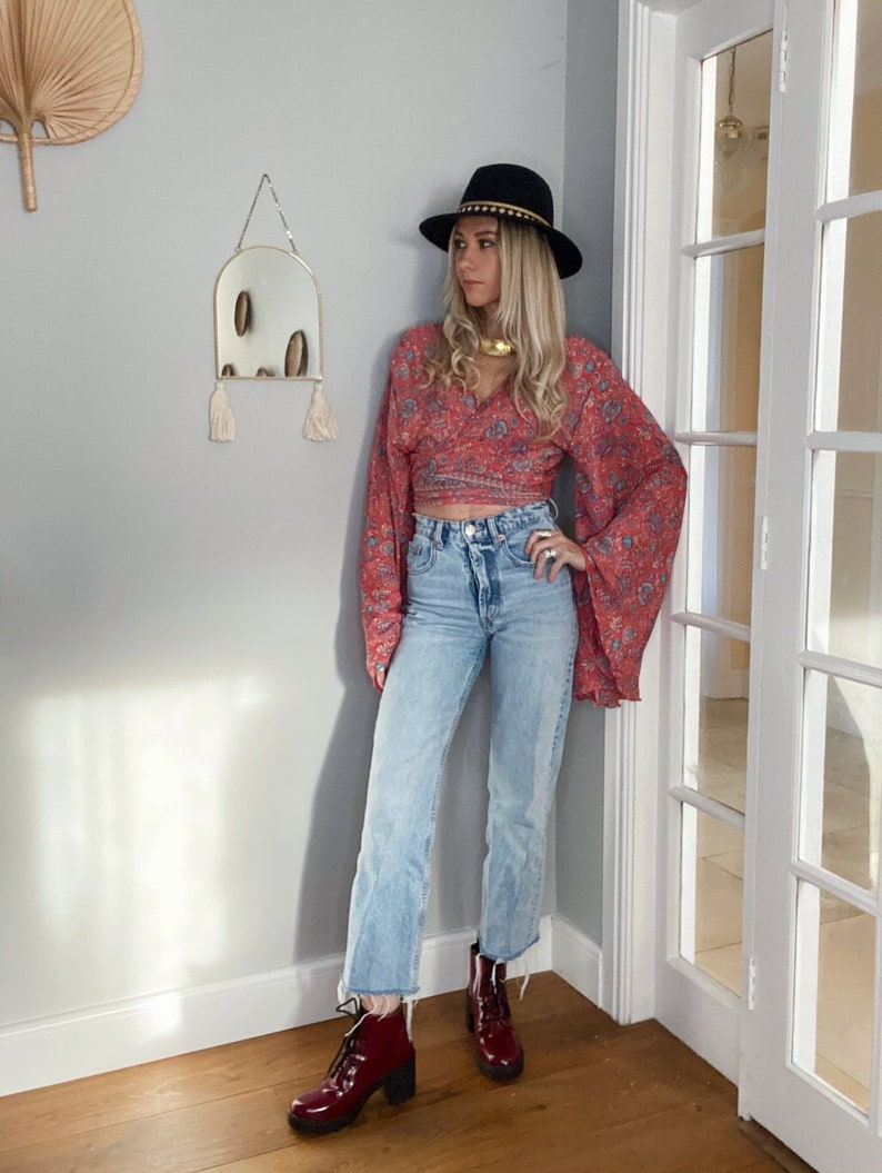May include: A woman wearing a red floral print wrap top with long bell sleeves, blue denim jeans, and burgundy ankle boots. She is wearing a black hat with a gold band and a gold necklace.