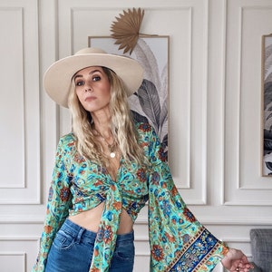 May include: A woman wearing a blue and green floral print top with a tie front and bell sleeves. She is also wearing a wide-brimmed white hat and blue jeans.