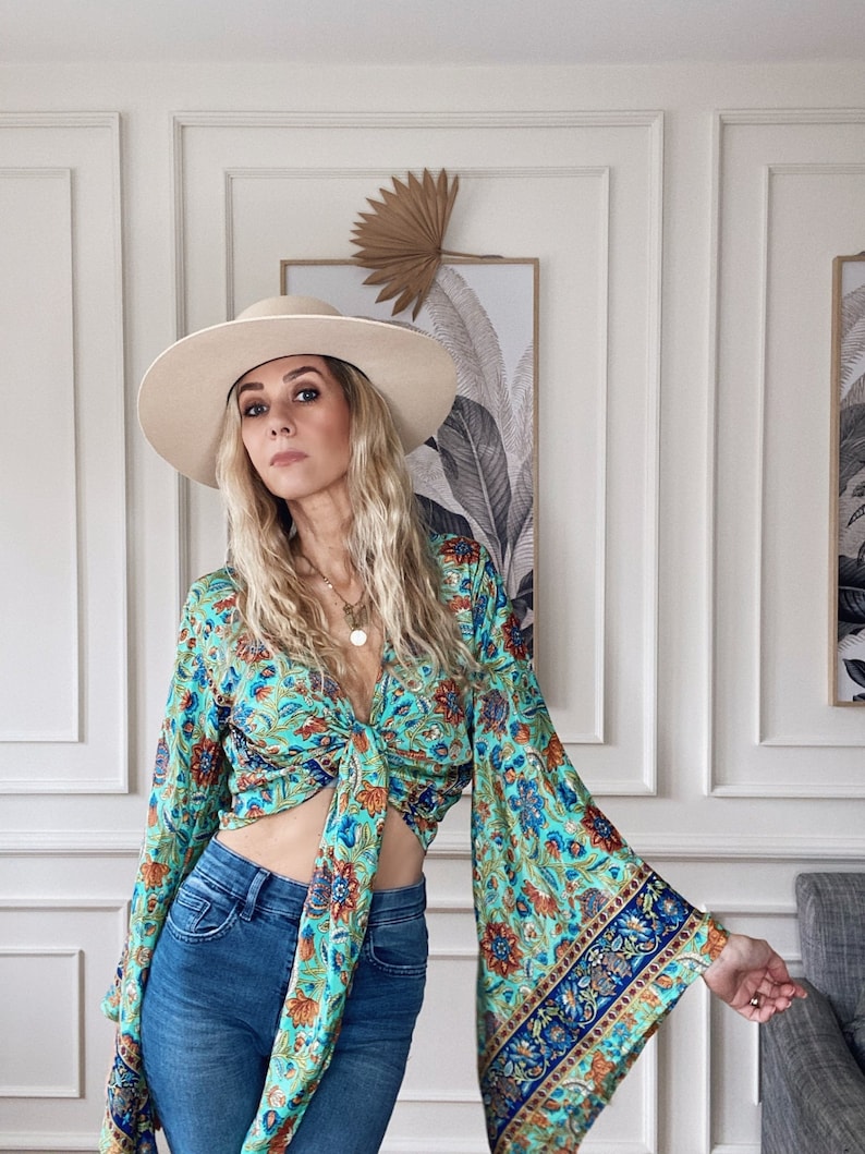May include: A woman wearing a blue and green floral print top with a tie front and bell sleeves. She is also wearing a wide-brimmed white hat and blue jeans.