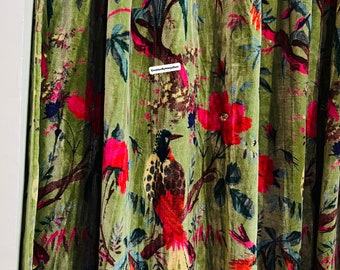 Pode incluir: Tecido de veludo verde com um padrão floral que apresenta flores cor-de-rosa, folhas verdes e pássaros coloridos. O tecido tem uma superfície texturizada, ligeiramente elevada.