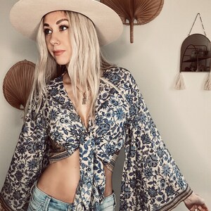 May include: A woman wearing a white hat, blue and white floral print top with a tie front, and blue jeans. The top has bell sleeves with gold trim.