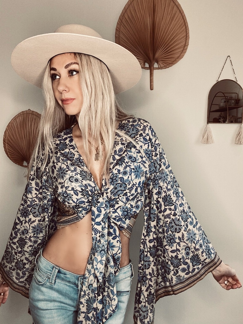 May include: A woman wearing a white hat, blue and white floral print top with a tie front, and blue jeans. The top has bell sleeves with gold trim.